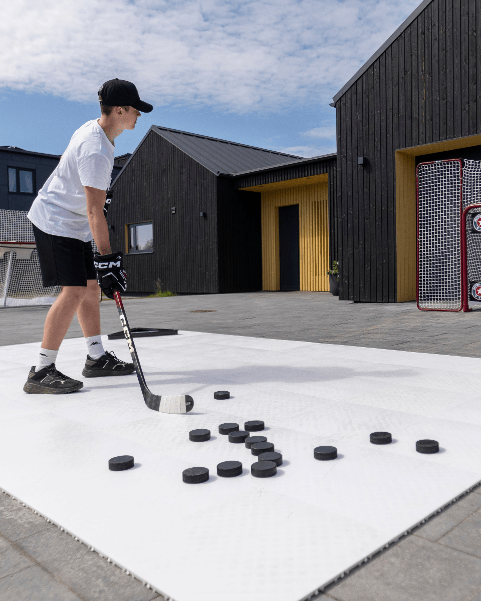 En ung hockeyspelare tränar på att skjuta puckar mot skottavlor på en utomhus isbana, omgiven av moderna byggnader.