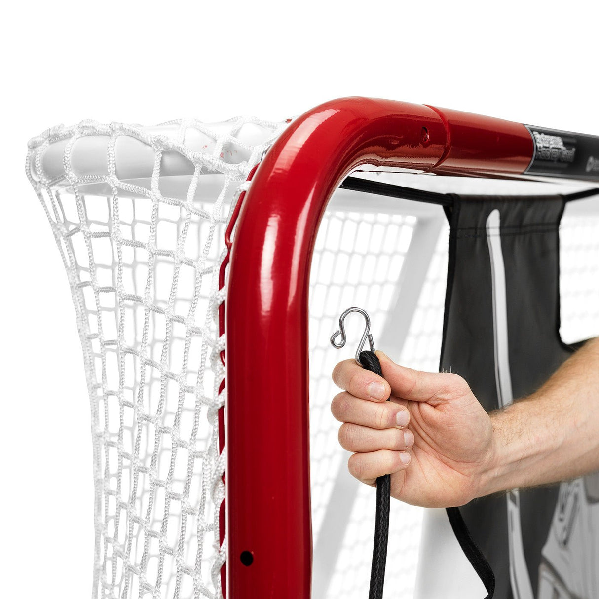 Närbild av en hand som fäster en krok på en målvaktsduk för hockey, med röd målvaktsram och nät i bakgrunden.
